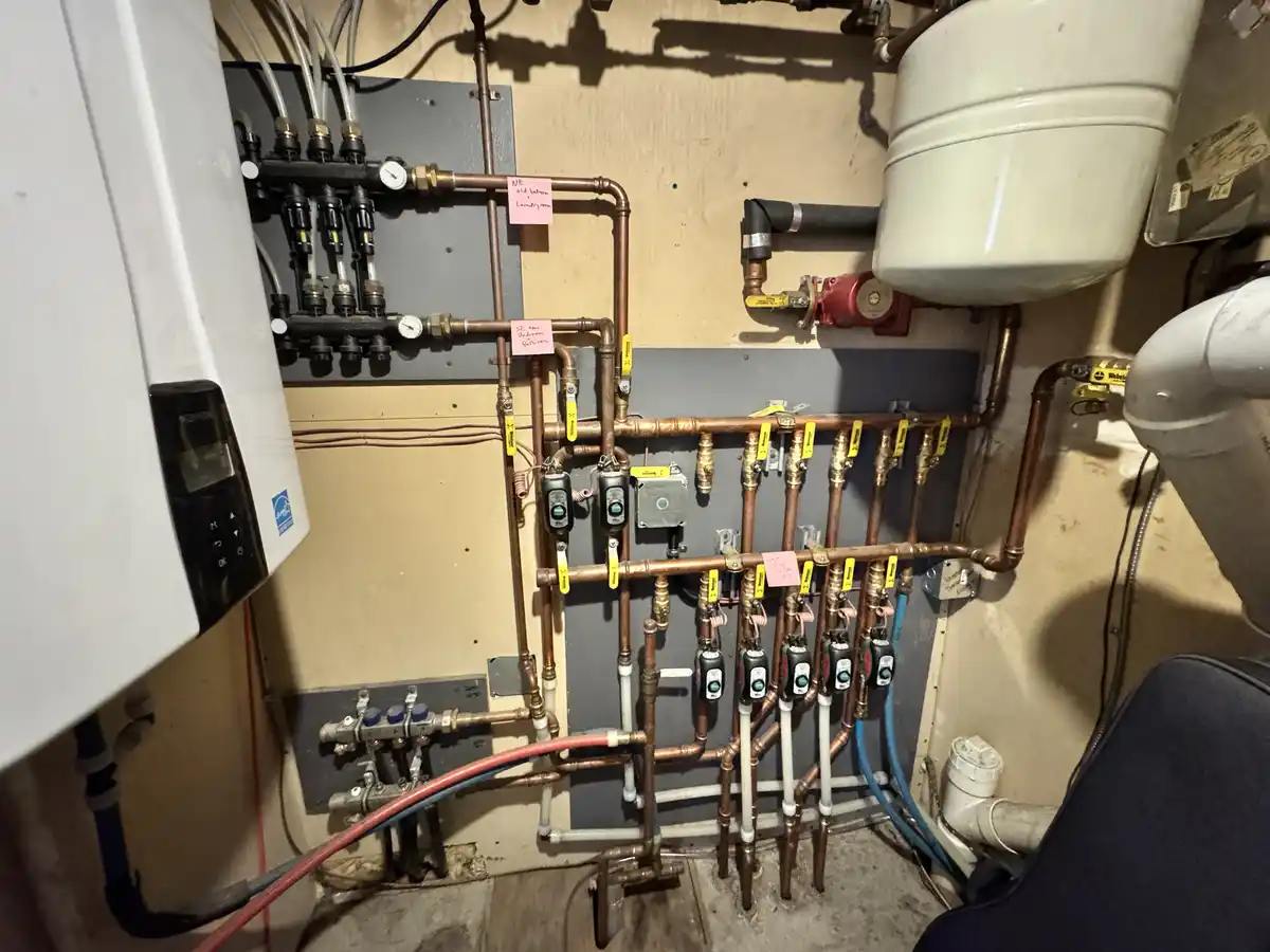 A complex network of copper pipes and valves in a utility room, with handwritten pink sticky notes labeling zones like 'NE…