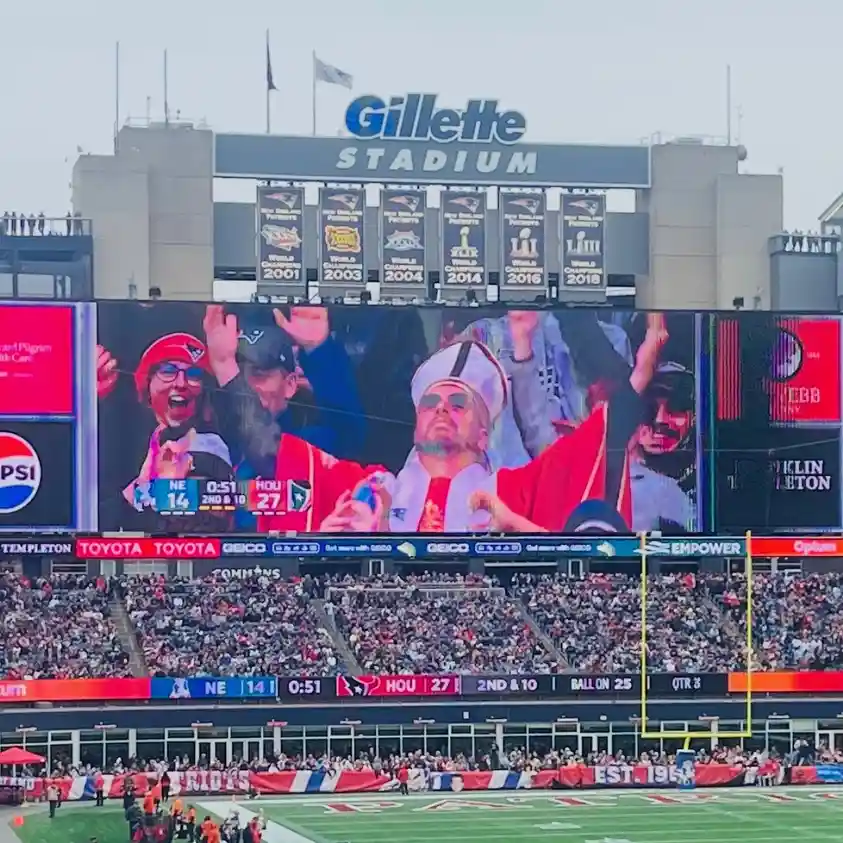 A large stadium scoreboard at Gillette Stadium shows a close-up of a man dressed in a red and white priest-like outfit with…