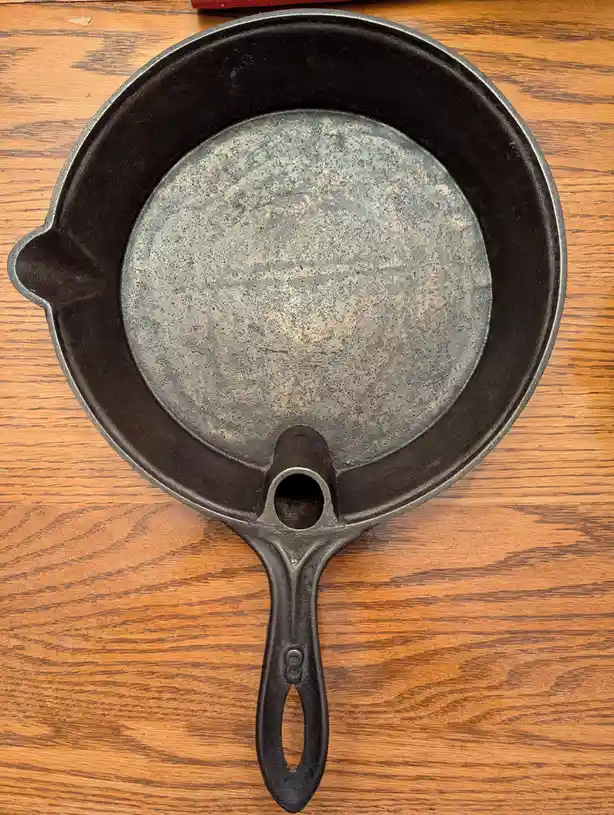 A top-down photo shows a well-used, black cast iron skillet resting on a wooden table. The skillet's surface is visibly…