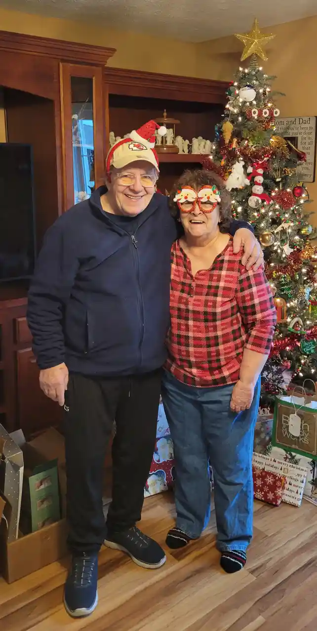 An older couple poses happily in front of a decorated Christmas tree. The man wears a baseball cap with a Santa hat on top…