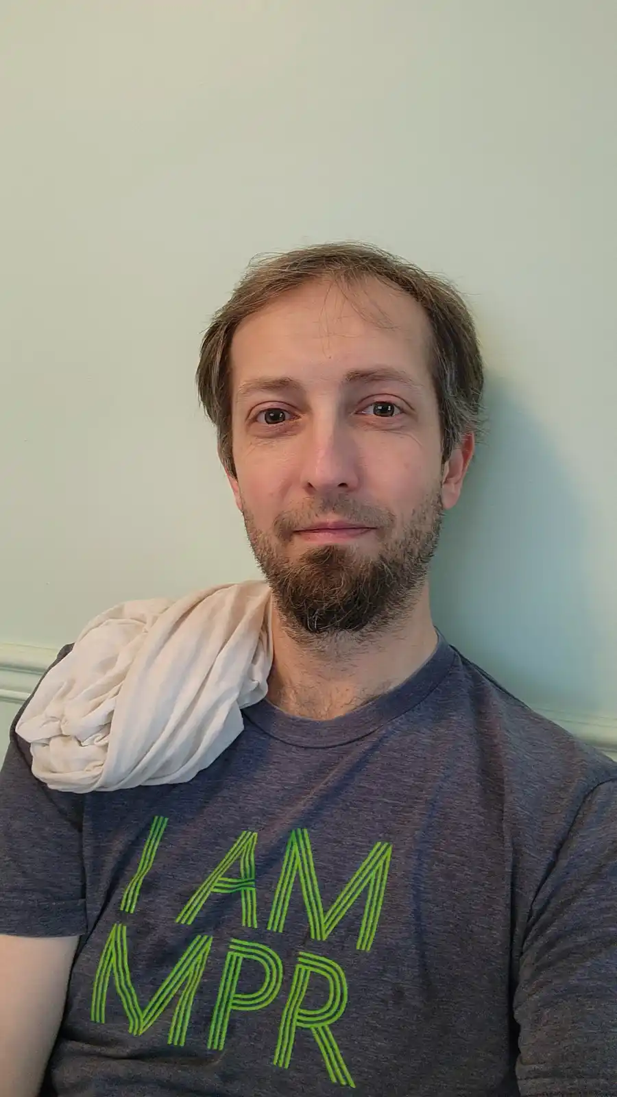 A man with a beard and short brown hair wears a gray t-shirt with bright green text that reads 'I AM MPR'. A white cloth is…