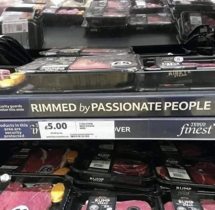 A supermarket meat aisle shelf with packaged steaks, where a black shelf label humorously reads 'RIMMED by PASSIONATE…