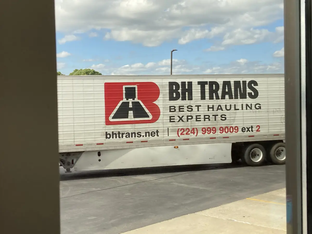 A white semi-trailer truck is parked outdoors under a partly cloudy sky, viewed through a window frame. The side of the…