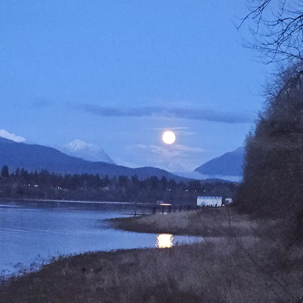 A serene twilight landscape featuring a full moon glowing over a calm body of water, with distant snow-capped mountains and…