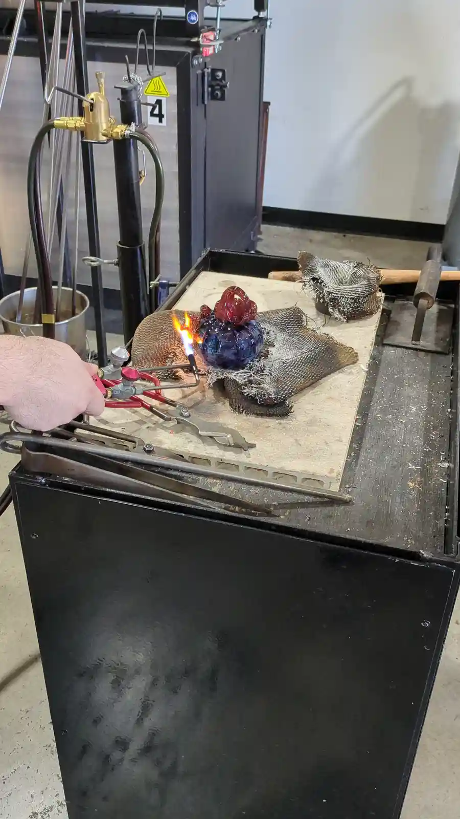 A person's hand holds a torch, heating a small, colorful glass sculpture on a workbench. The sculpture has a blue base and…