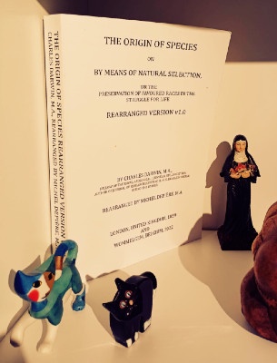 A book titled 'The Origin of Species' with 'REARRANGED VERSION v2.6' on the cover, flanked by a small blue dog figurine and…