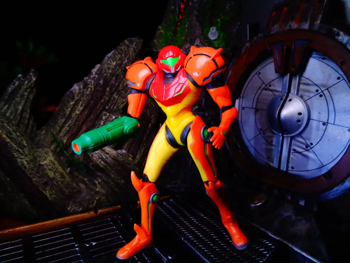 A Samus Aran action figure in her iconic orange and yellow Power Suit stands in a sci-fi diorama, holding a green weapon…