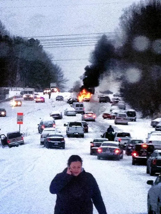 A woman in a dark jacket walks through deep snow on a road, holding a phone to her ear, while behind her, a major traffic…