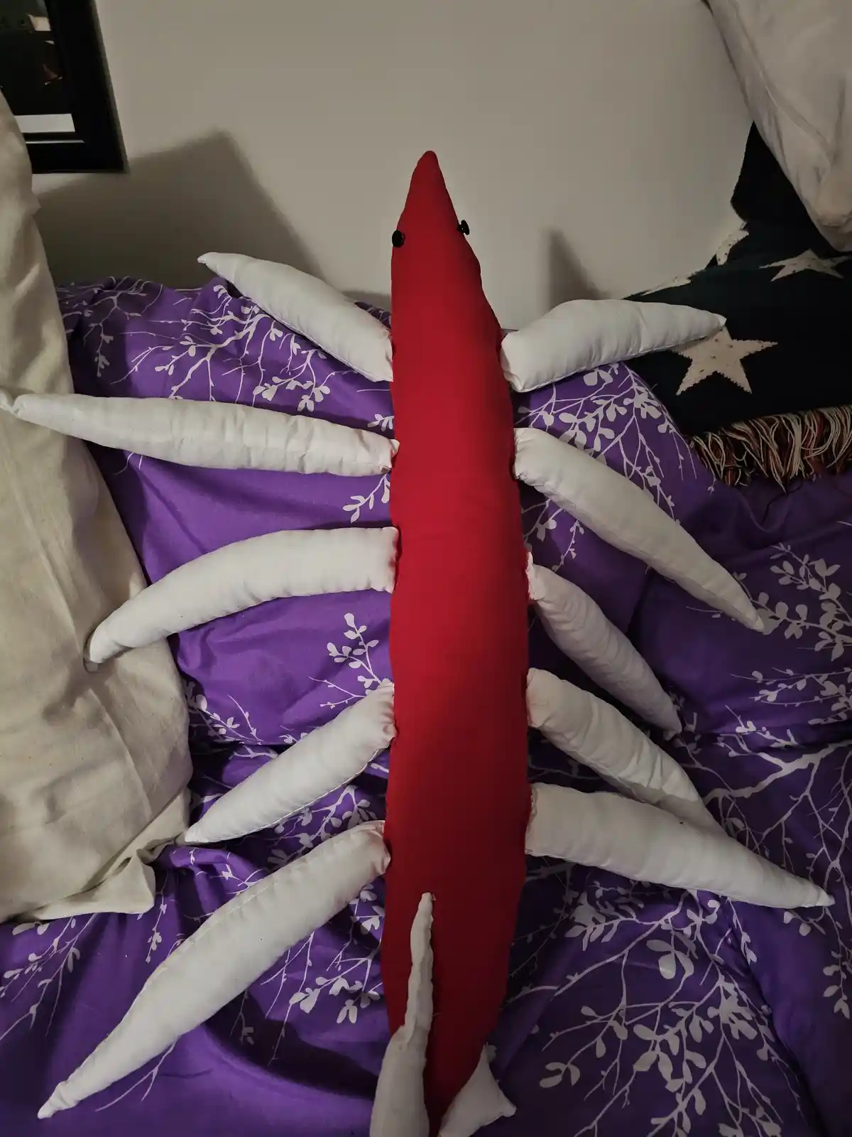 A red, elongated, pillow-like creature with black button eyes sits upright on a purple bedspread with white vine patterns…