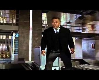 A man in a black suit and tie stands in a modern, industrial-looking building with metal railings and concrete walls. He has…