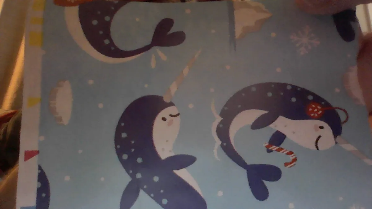 A close-up photo of a children's fabric or wrapping paper featuring cartoon narwhals in a blue, snowy underwater scene. One…