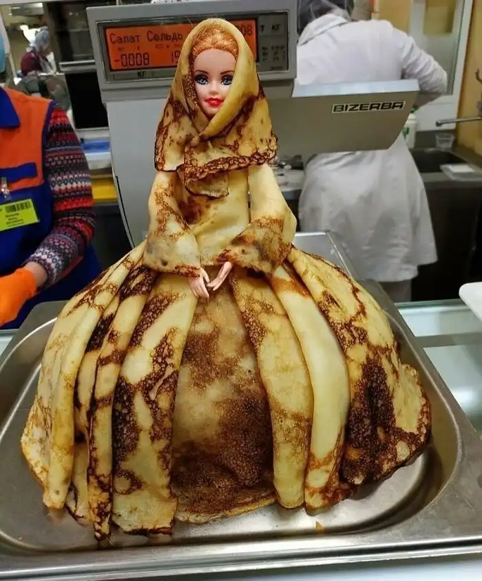 A doll with a blonde wig and red lips is dressed in a voluminous, multi-layered dress made entirely of golden-brown…