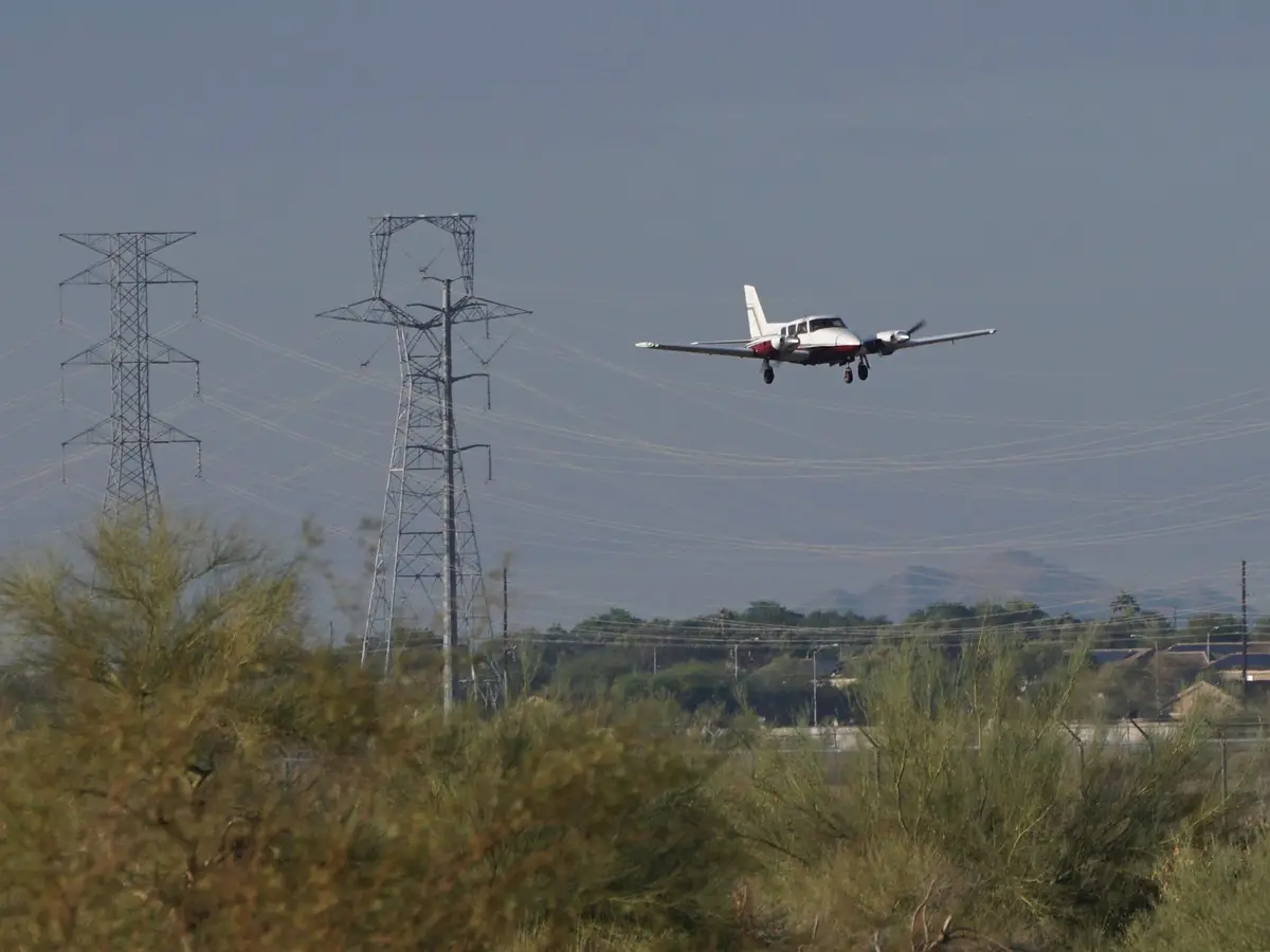 A small white and red propeller airplane is flying low with landing gear down, positioned directly between two tall…