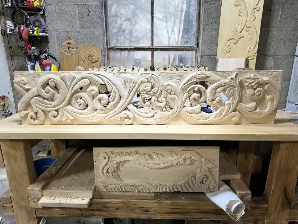 A long, ornately carved wooden piece with swirling, baroque-style patterns sits on a workbench in a workshop, with another…