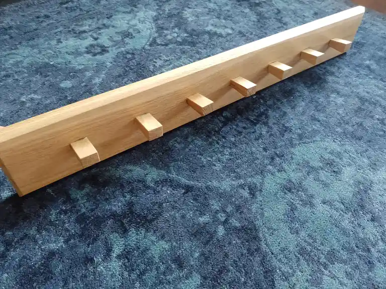 A light-colored wooden coat rack with five small square pegs lies on a blue patterned carpet. The overlaid text reads 'i…