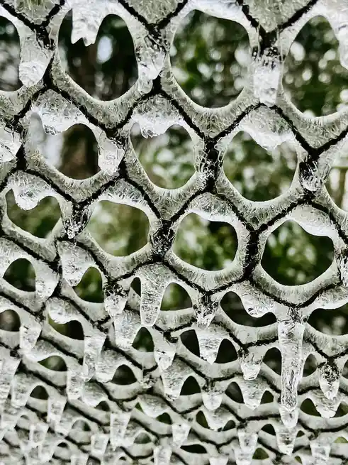 A close-up photo shows a black mesh fence covered in thick, clear ice formations, with icicles hanging from the edges of the…