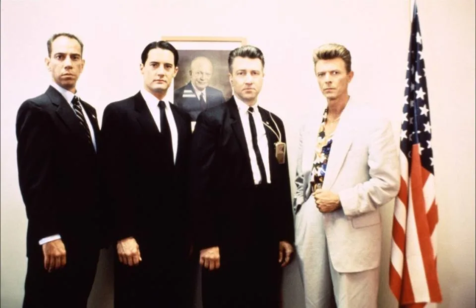 Four men stand in a formal office setting, three in dark suits and one in a light-colored suit with a patterned shirt…