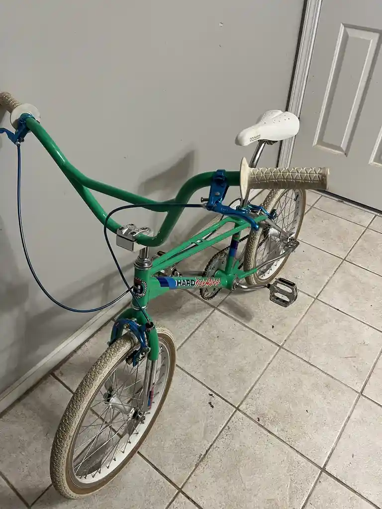 A green BMX bike with blue handlebars and a white saddle is leaning against a wall on a tiled floor. The bike's frame has…