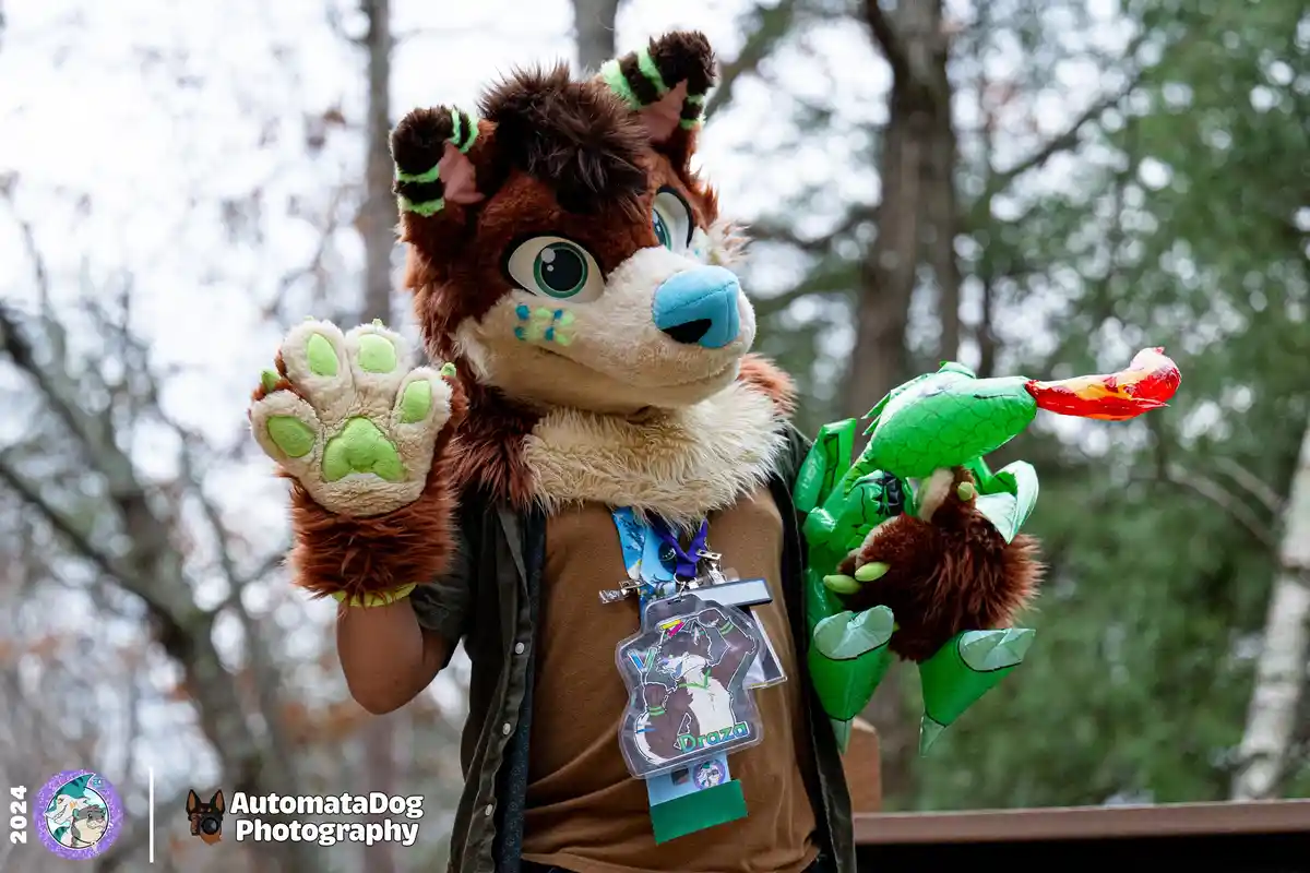 A person in a brown and cream fursuit with green accents and large blue eyes holds up a green paw and a green dragon plushie…
