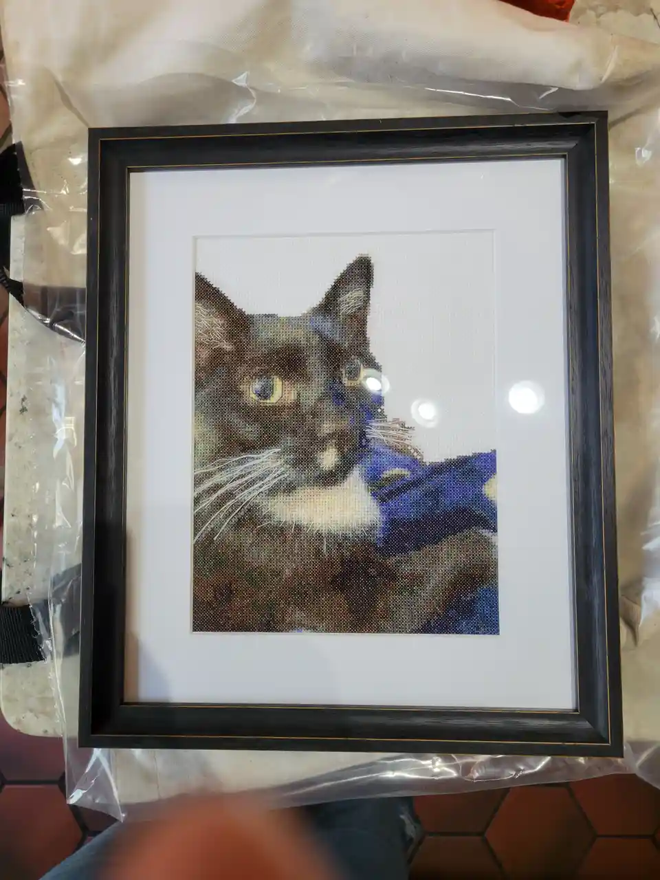 A framed cross-stitch portrait of a cat with brown, black, and white fur, yellow eyes, and a blue collar, displayed on a…