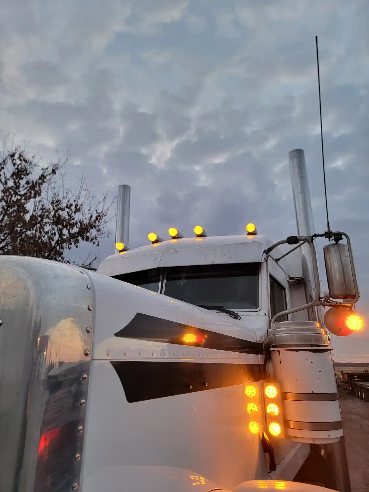 A low-angle photo of a white semi-truck cab at dusk, with multiple glowing amber lights on the roof and side, against a…