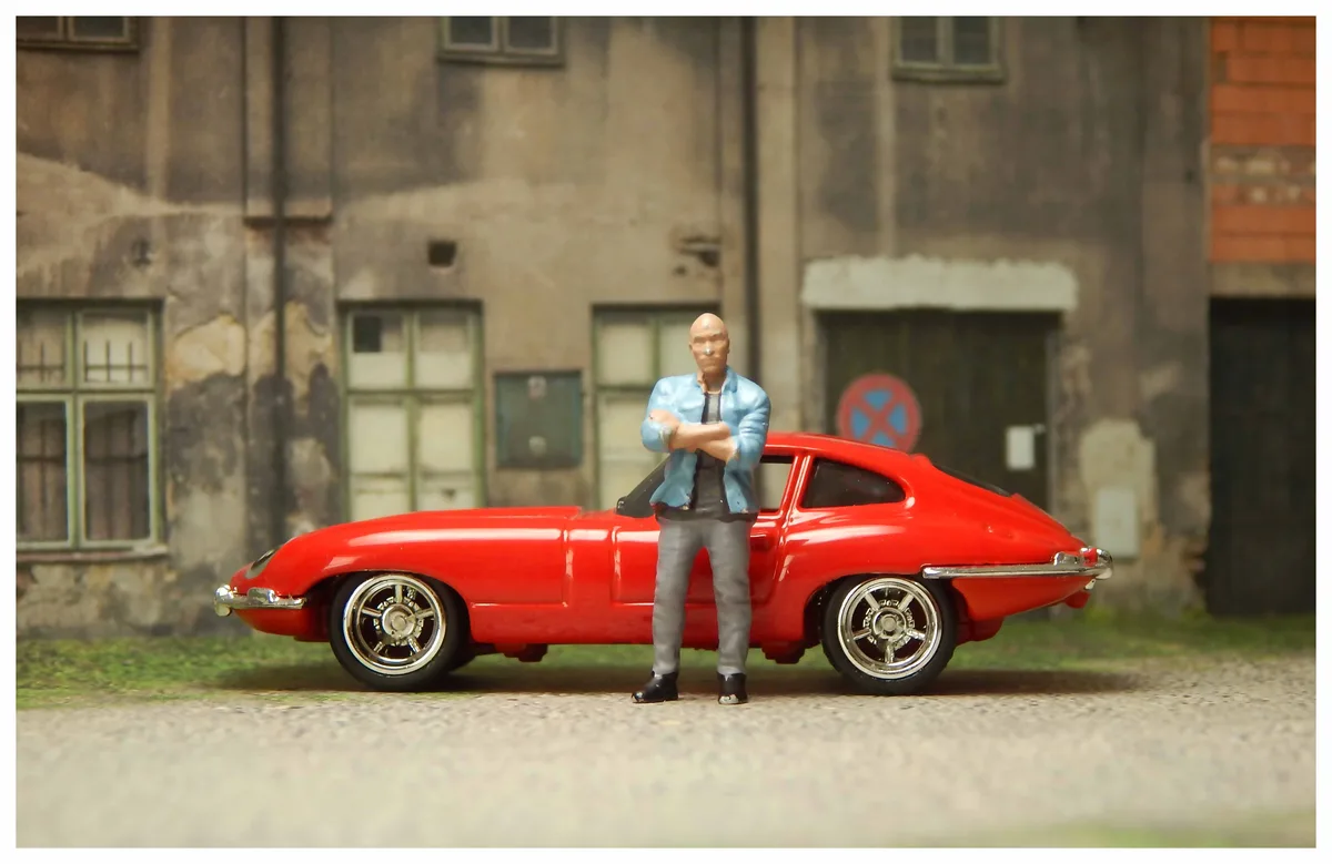A toy figurine of a bald man in a blue jacket and grey pants stands with arms crossed beside a red toy Jaguar E-Type sports…