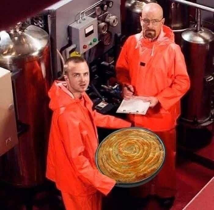 Two men in orange jumpsuits stand in a lab-like setting with industrial equipment. The man in the foreground holds a large…