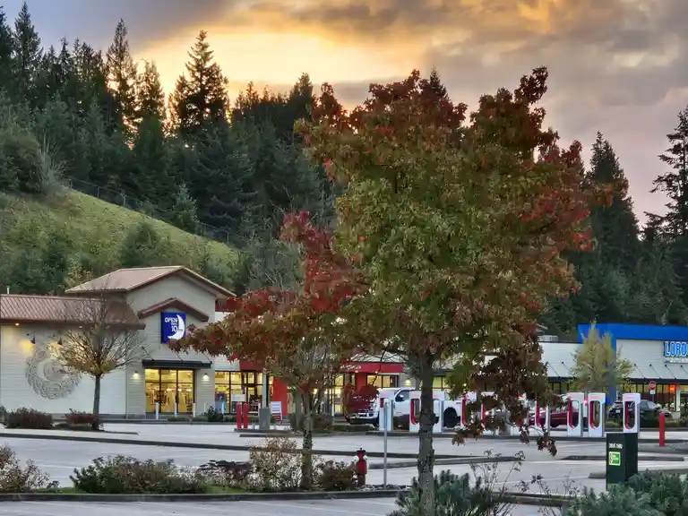 A commercial strip mall at dusk with a prominent sign reading 'OPEN UNTIL 10PM' on a building, surrounded by trees with…
