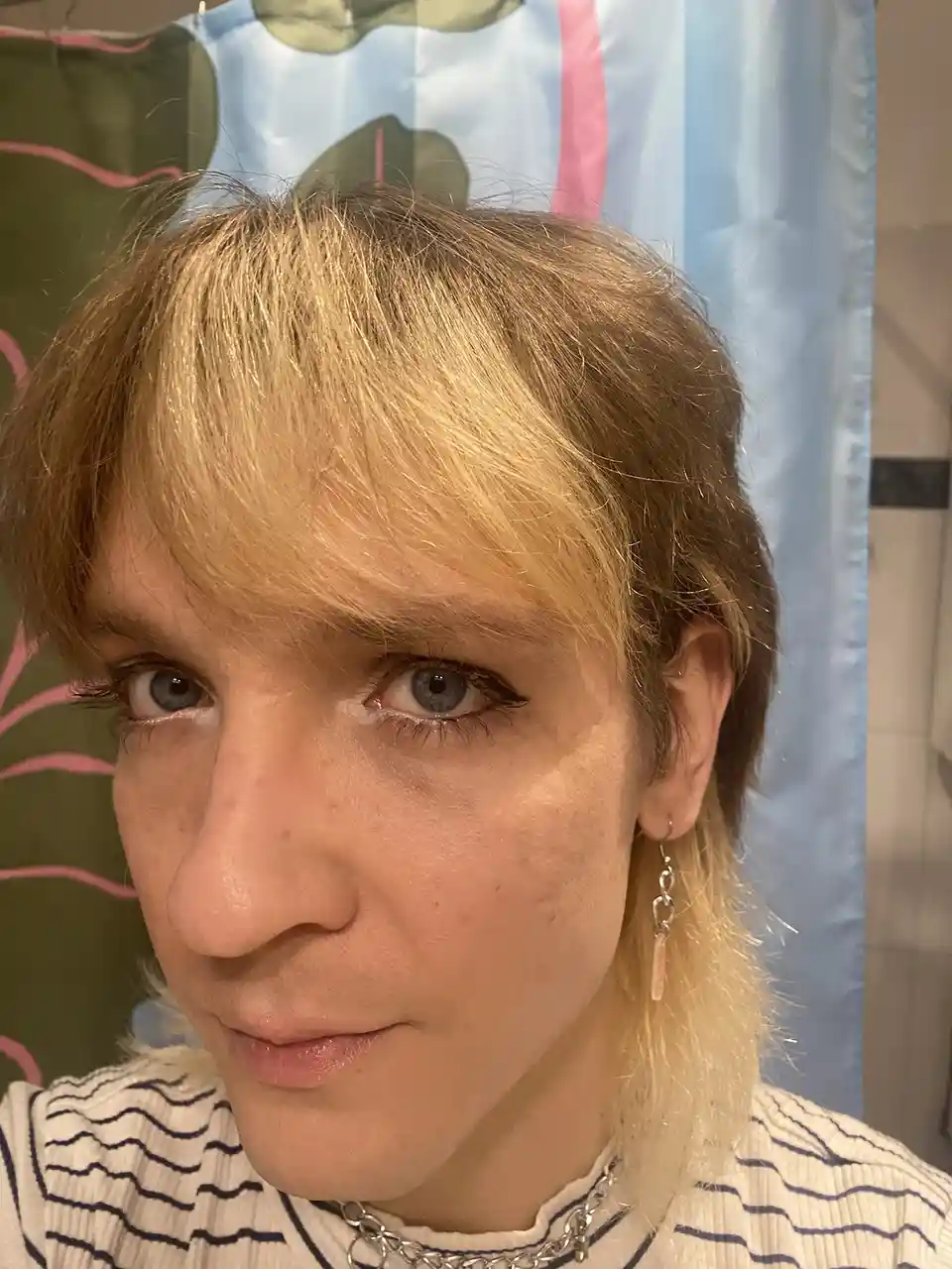 A person with blonde hair, blue eyes, and makeup looks at the camera with a slight smirk, wearing a striped shirt, a chain…