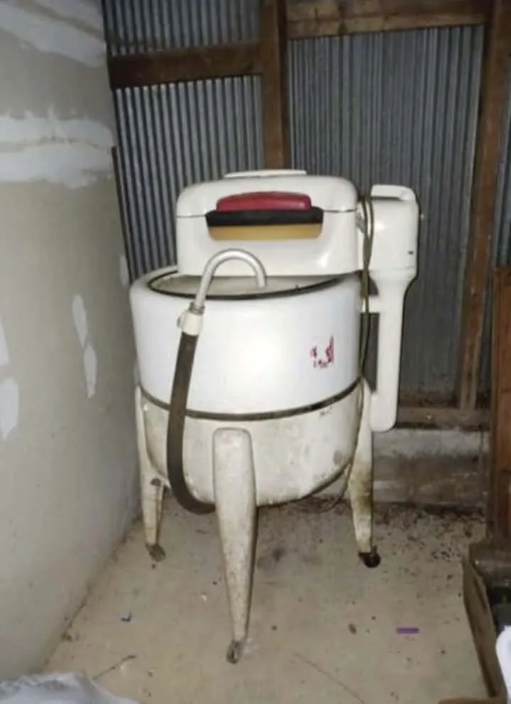 A dirty, white, freestanding washing machine with a red handle and hose, standing in a dusty, unfinished room with…