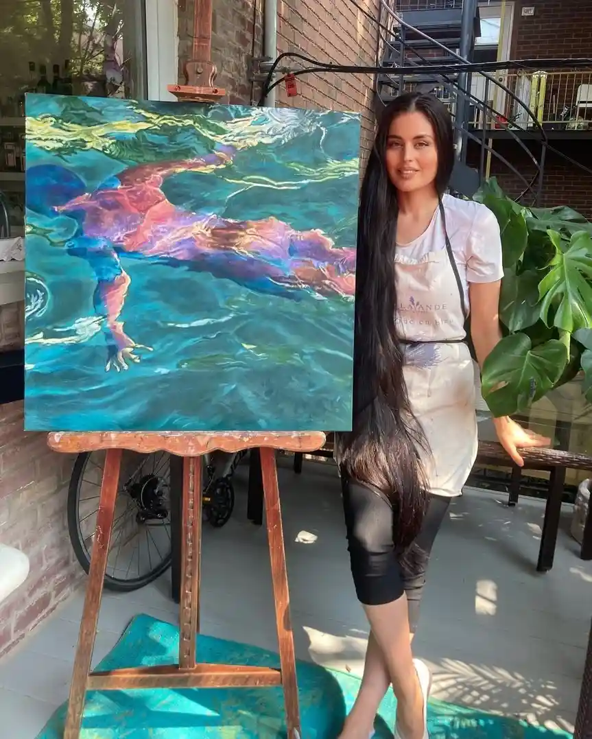 A woman with long dark hair stands beside a large painting on an easel, wearing a paint-splattered apron. The painting…