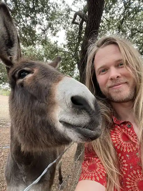 A man with long blonde hair and a beard, wearing a red patterned shirt, takes a selfie with a donkey. The donkey's head is…
