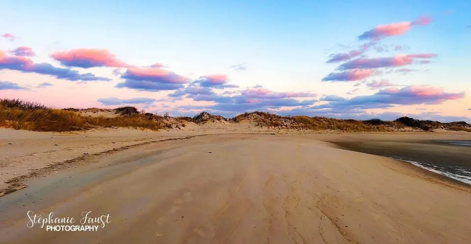 A serene beach scene at sunset with pink and blue clouds over sand dunes and wet sand leading to the ocean, overlaid with…