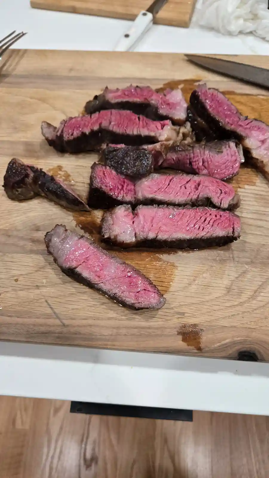 sliced medium-rare steak with pink center and seared edges rests on a wooden cutting board, with kitchen knives and a fork…
