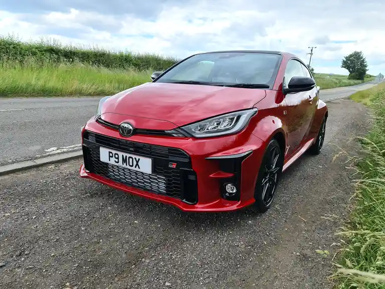 A red Toyota GR Yaris parked on a gravel shoulder next to a road, with green hedgerows and a cloudy sky in the background…