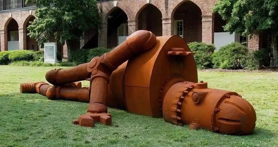 A large, rusted metal sculpture of a human figure lying on its back on a grassy lawn in front of a brick building with…
