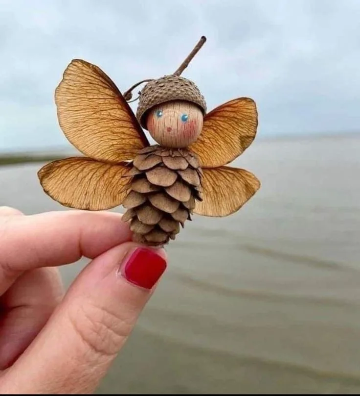 A hand with red nail polish holds a tiny, handcrafted figurine made from natural materials: a pinecone body, acorn cap head…