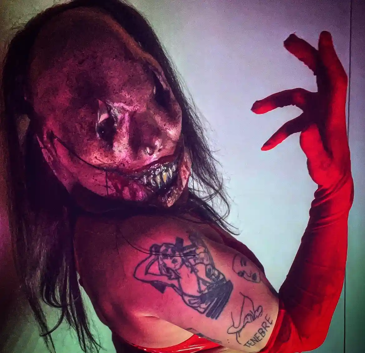 A person with long dark hair wears a grotesque, decaying monster mask with sharp teeth and red lighting, raising a…