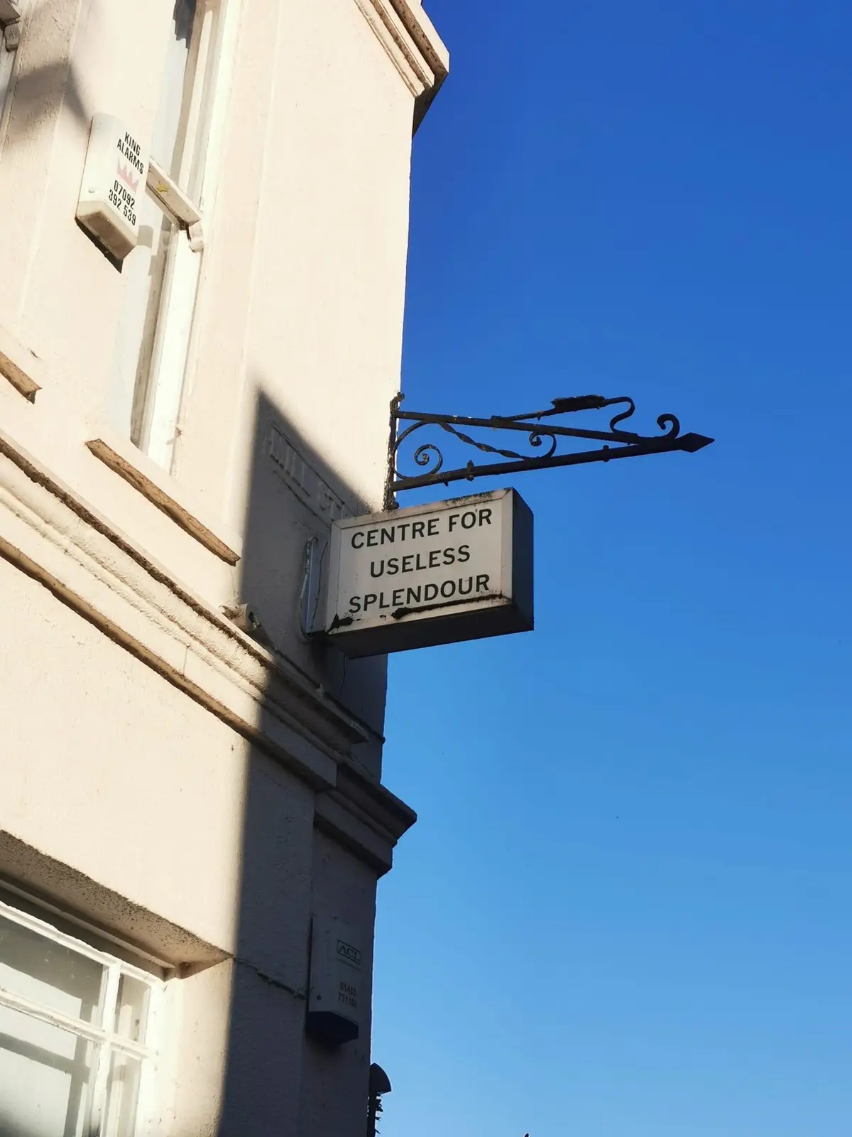 A white rectangular sign with black text reading 'CENTRE FOR USELESS SPLENDOUR' is mounted on an ornate black metal bracket…