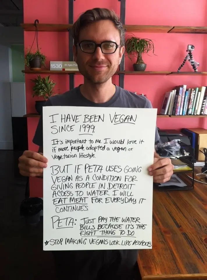 A man with glasses holds a handwritten sign stating he's been vegan since 1999 and supports veganism, but will eat meat…