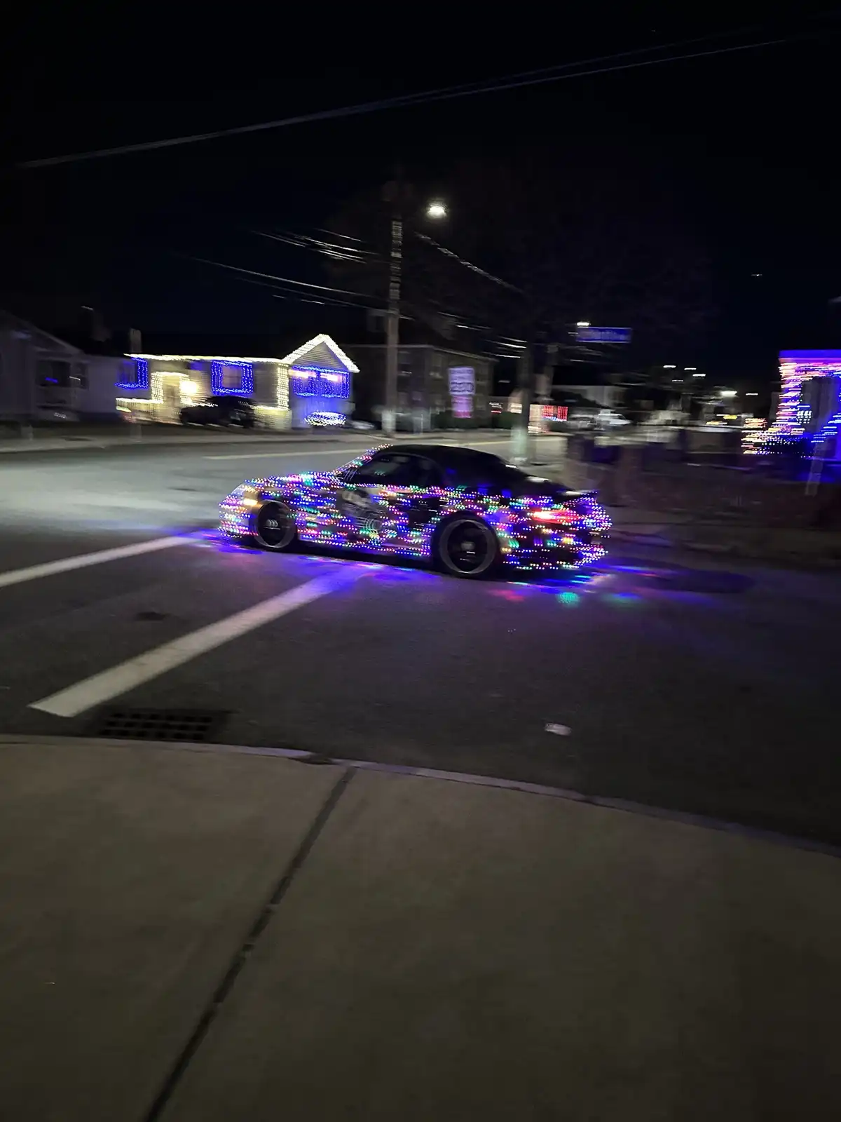 A black sports car covered in blinking multicolored LED lights drives down a residential street at night, casting colorful…