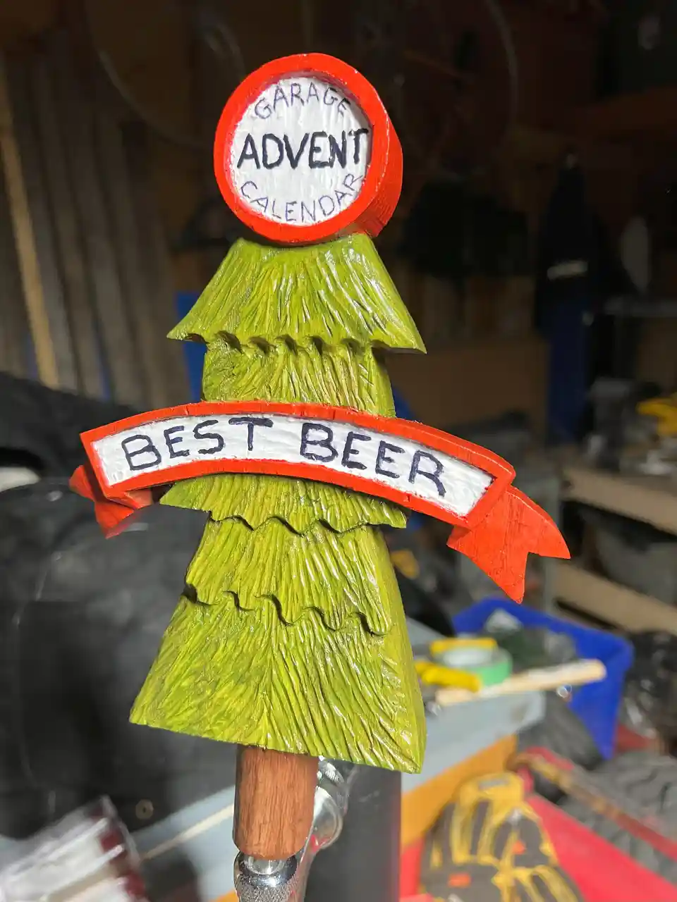 A hand-carved green Christmas tree-shaped beer tap handle with a red circular top reading 'GARAGE ADVENT CALENDAR' and a red…