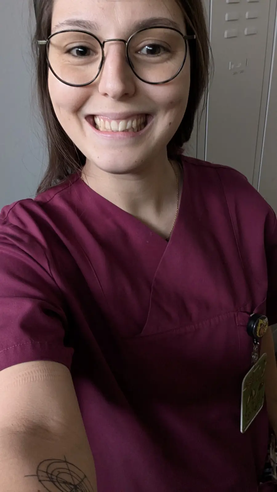 A smiling woman in maroon scrubs and round glasses takes a selfie, showing her teeth with a proud expression. She wears a…