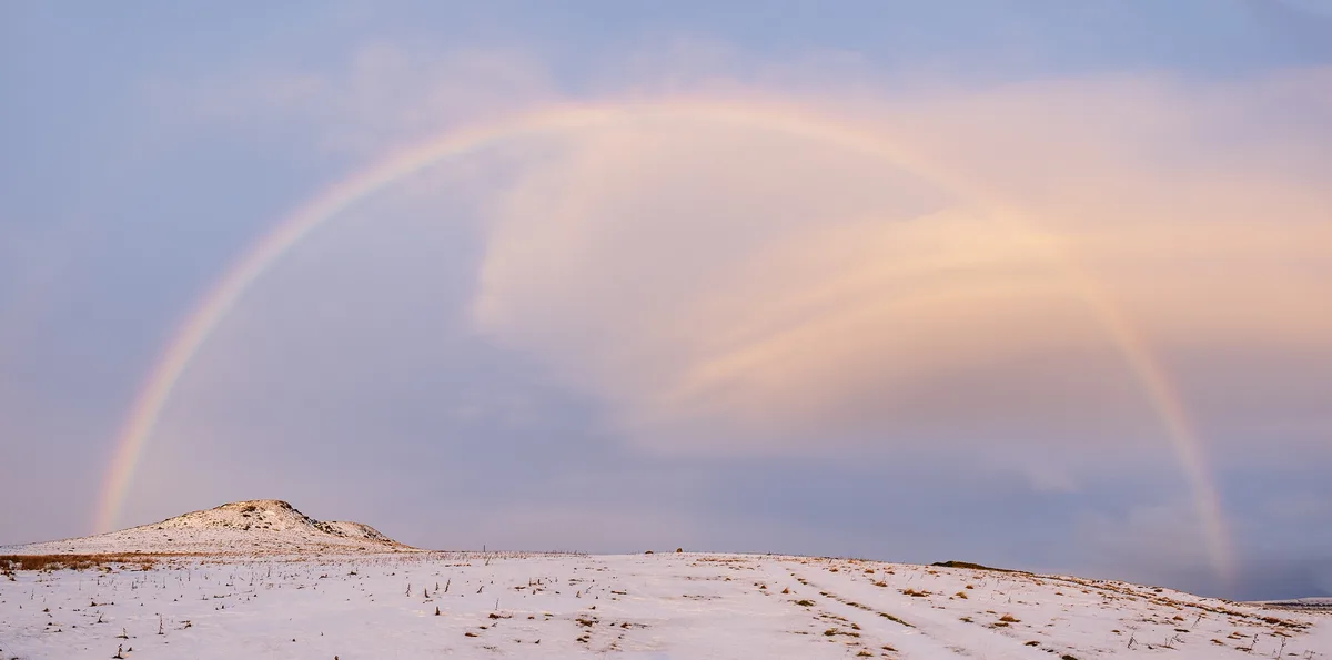 A wide landscape photo shows a snowy, hilly terrain under a pale blue sky with soft clouds. A faint, pale rainbow arcs…