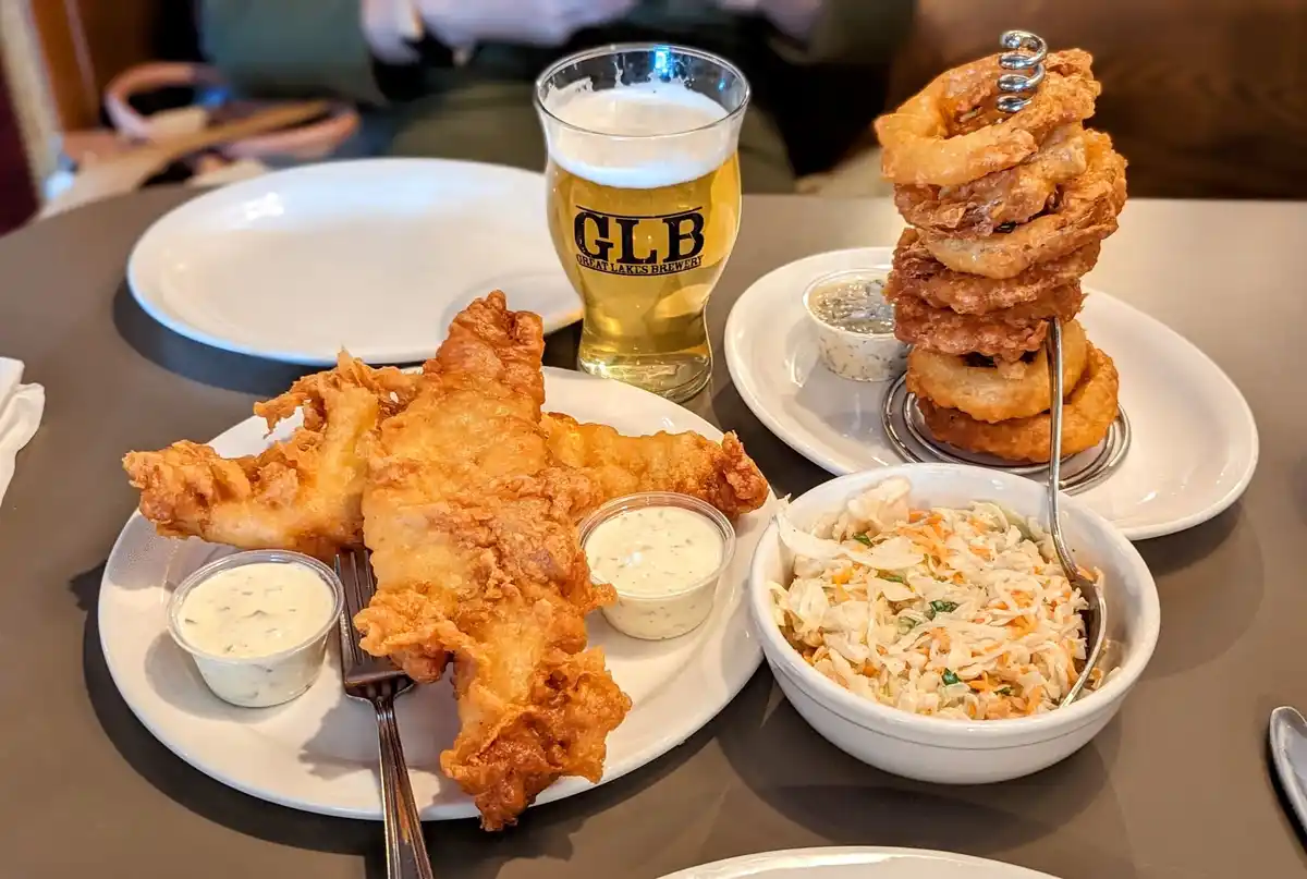 A table setting features a plate of golden-brown fried fish with tartar sauce, a towering stack of fried onion rings on a…