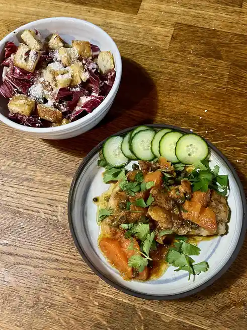 Two dishes sit on a wooden table: a bowl of red radicchio salad topped with croutons and grated cheese, and a plate with a…