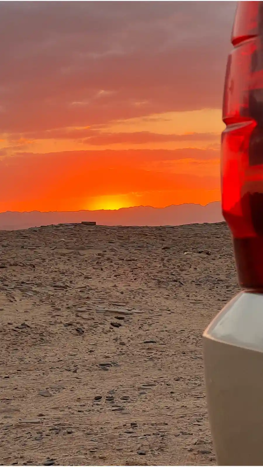 A photo taken from inside a vehicle shows a stunning orange and red sunset over a rocky desert landscape with distant…