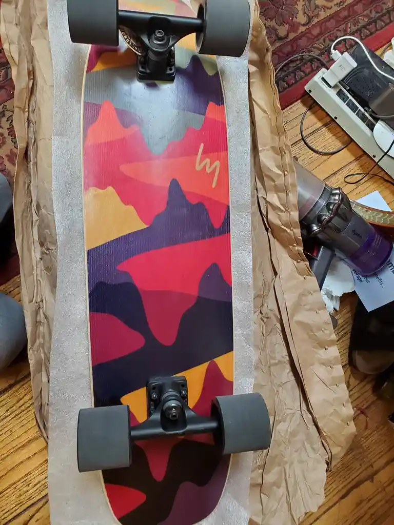 A longboard with a colorful mountain-patterned deck is shown lying on brown packing paper on a wooden floor, surrounded by…