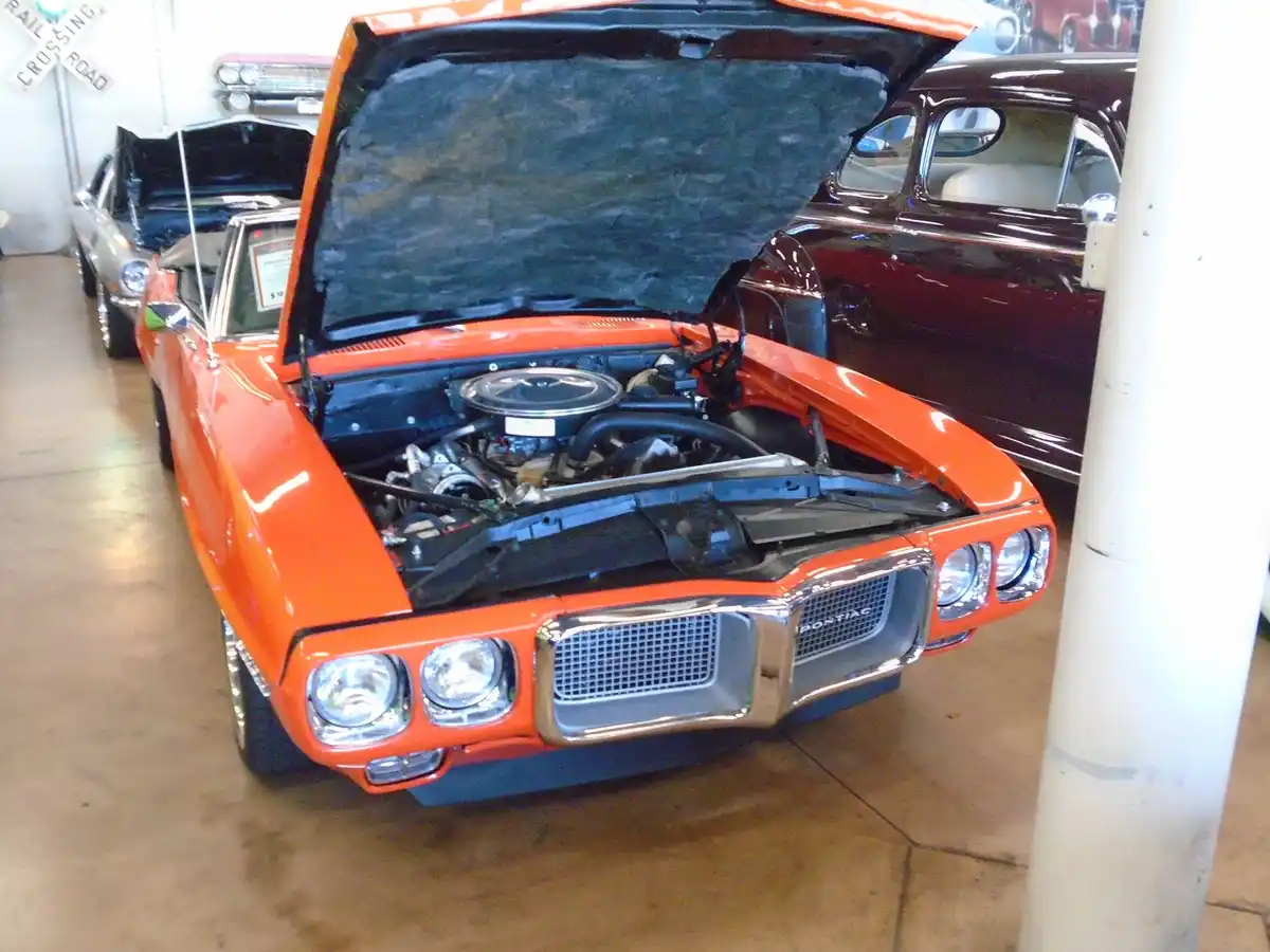 An orange classic Pontiac GTO with its hood open, revealing the engine, is parked indoors among other vintage cars. The…
