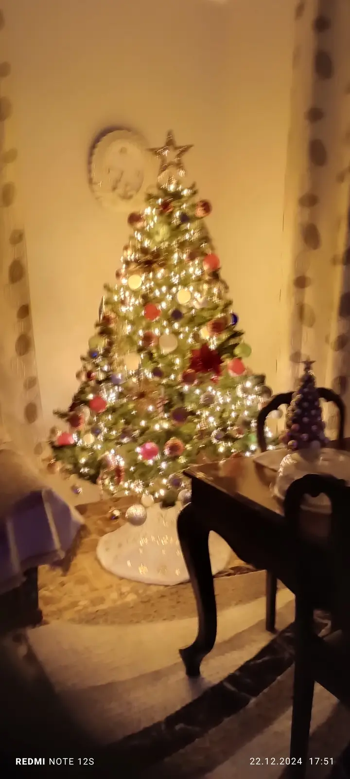 A blurry indoor photo shows a brightly lit, decorated Christmas tree with colorful ornaments and a star on top, standing…